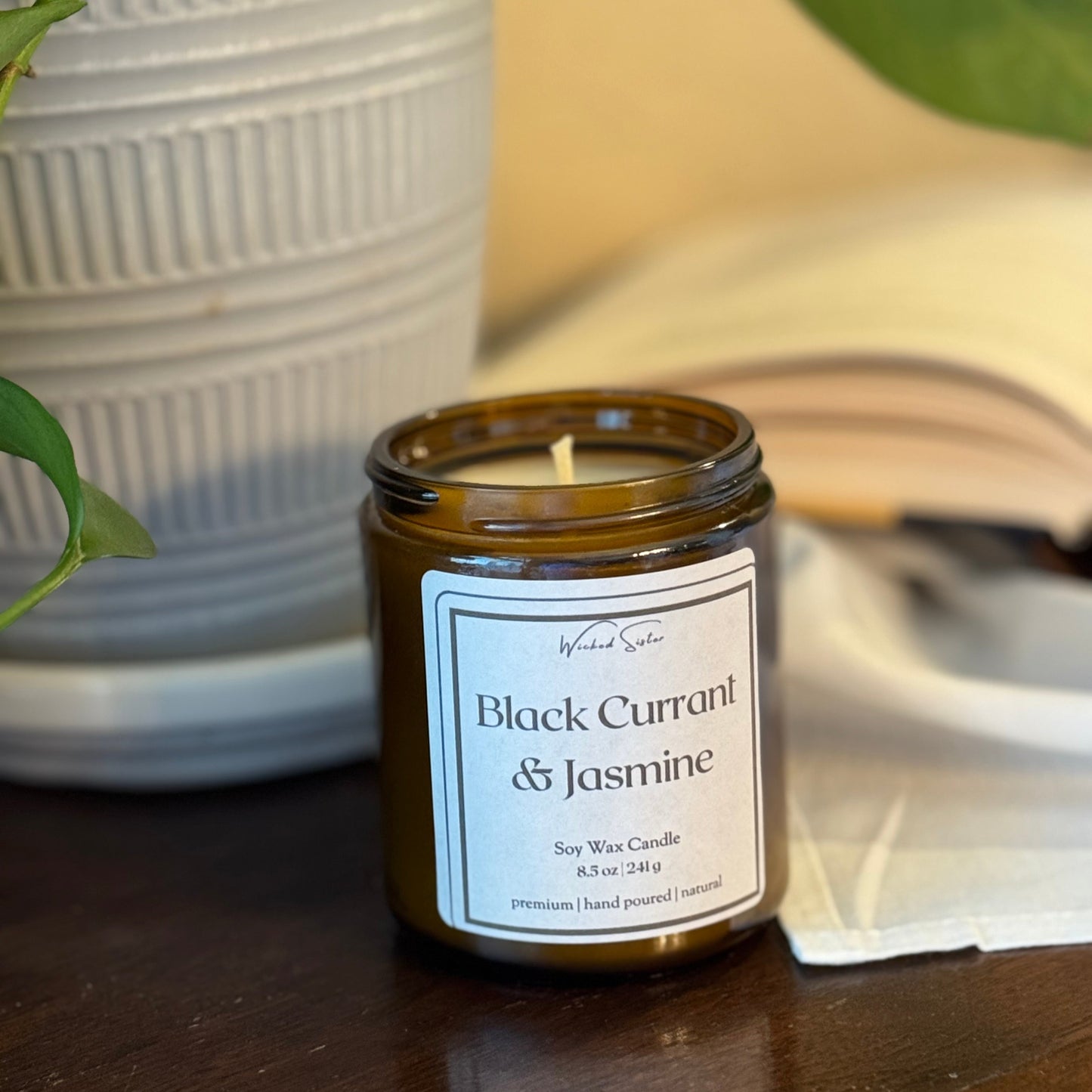 Candle labeled 'Black Currant & Jasmine' on a wooden surface with a plant and book in the background.