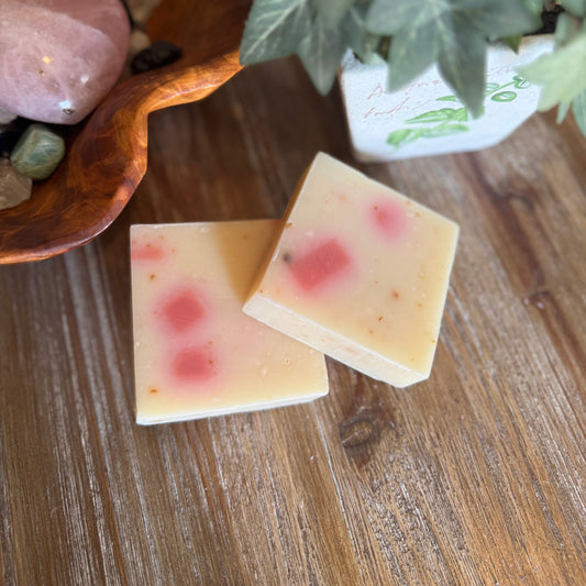 Two soap bars with pink specks on a wooden surface next to a plant and bowl of stones.