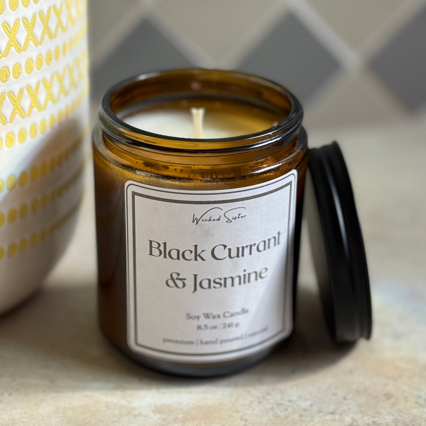 Candle jar labeled 'Black Currant & Jasmine' on a textured surface with a patterned background
