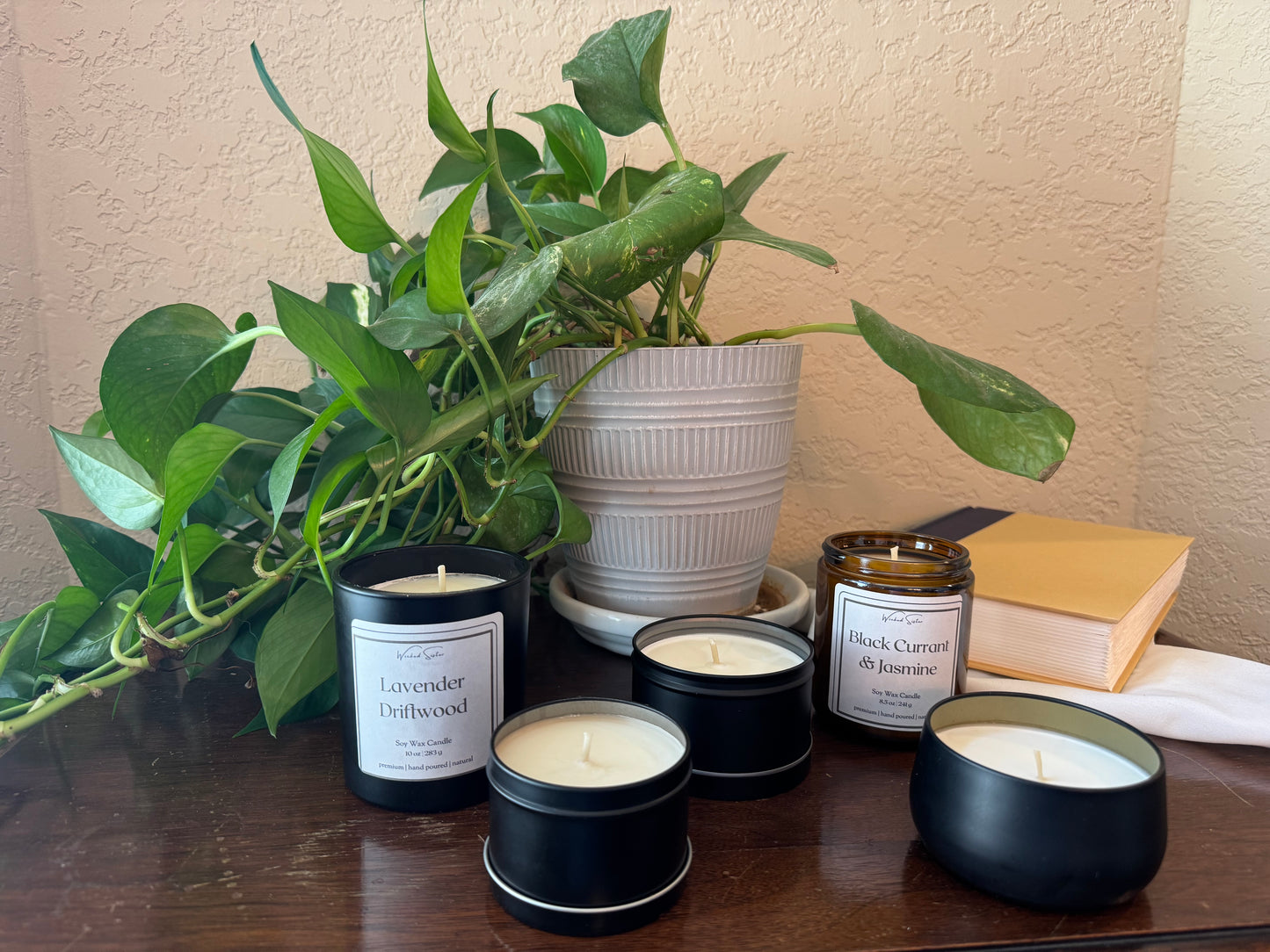 Collection of candles with visible labels on a table next to a plant and books.