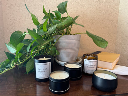 Collection of candles with visible labels on a table next to a plant and books.