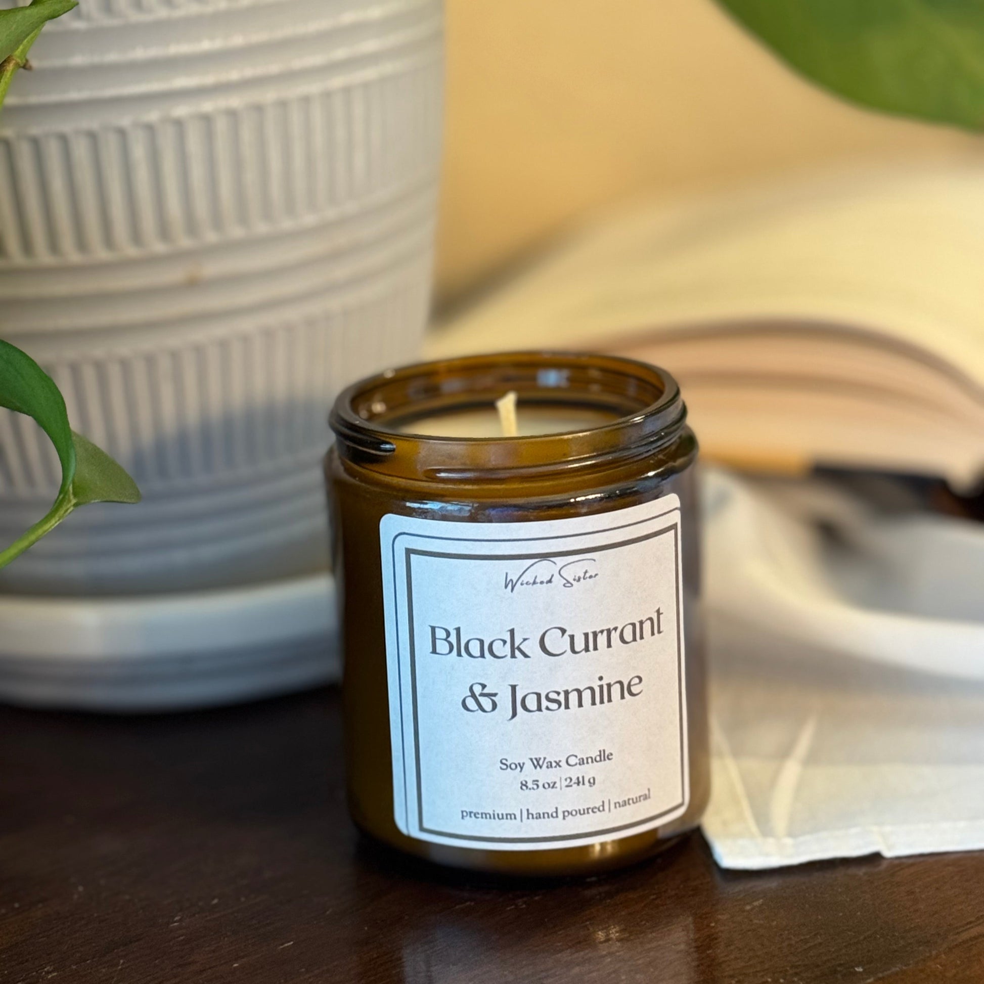 Candle labeled 'Black Currant & Jasmine' on a wooden surface with a plant and book in the background.