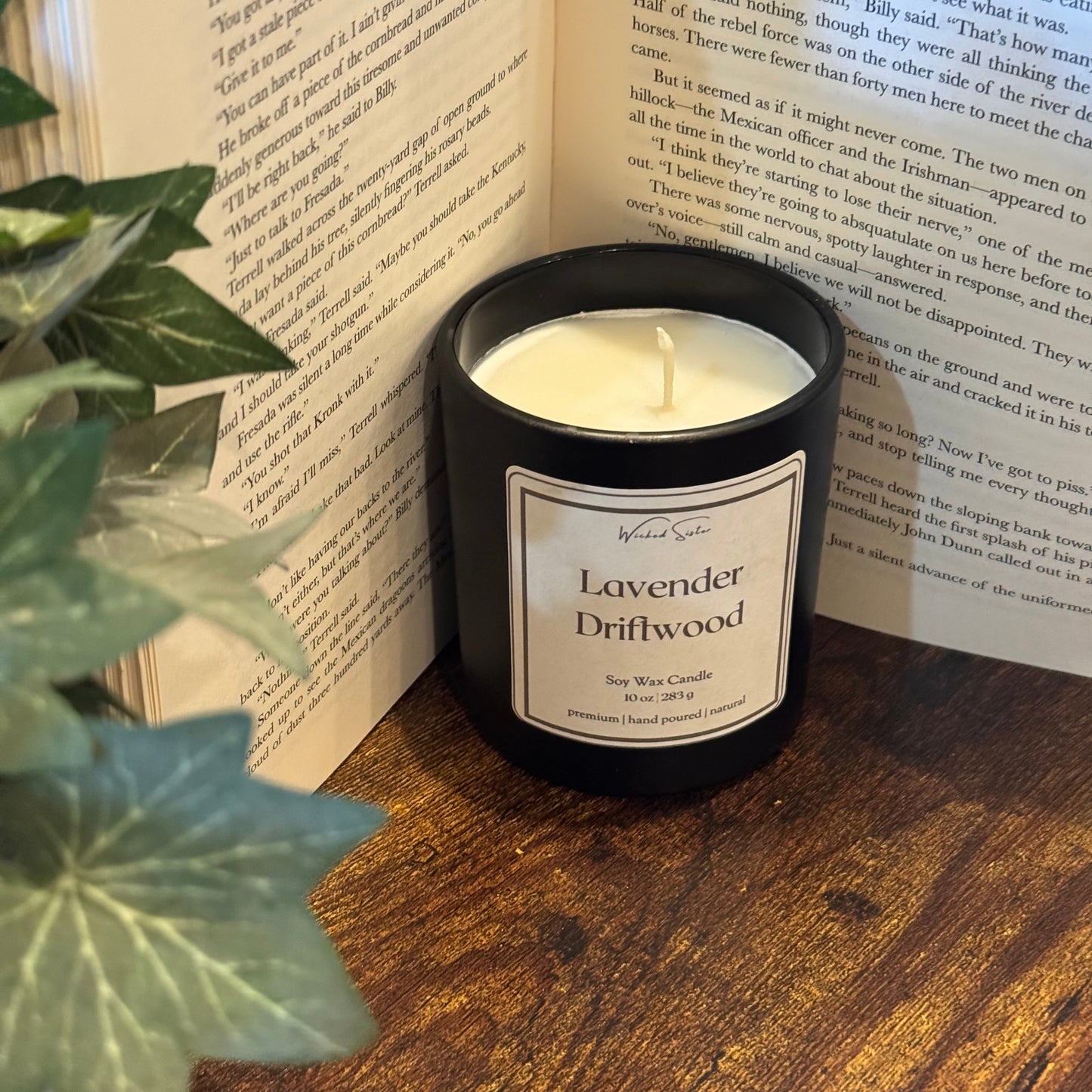 Candle labeled 'Lavender Driftwood' on an open book with a plant in the foreground.