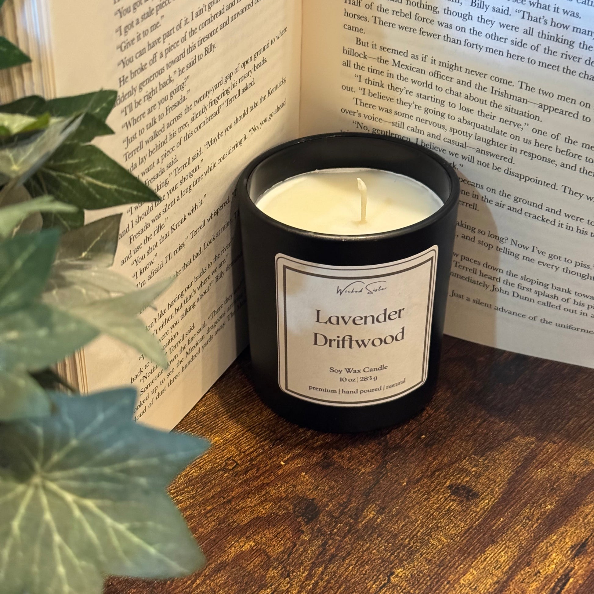 Candle labeled 'Lavender Driftwood' on an open book with a plant in the foreground.